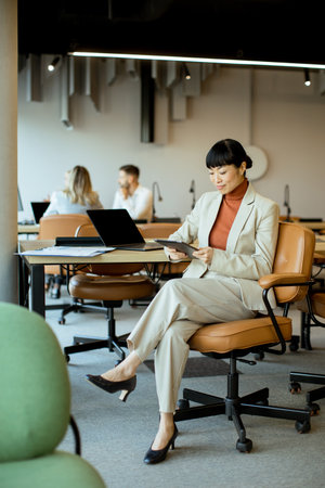 A woman in a beige suit sits at her desk, focused on reviewing documents on a tablet. The contemporary office features other workers engaged in various activities in the background.の写真素材