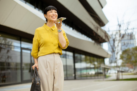 A woman is smiling while speaking on her smartphone outside a contemporary building in the city. She is dressed in a yellow blouse and beige pants, holding a black bag.の写真素材