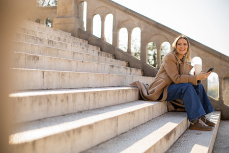 A cheerful woman dressed in a long coat and jeans sits comfortably on stone steps. She smiles while holding her phone, enjoying a sunny day at a beautiful park.の写真素材