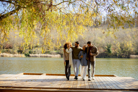 Four friends walk happily along a wooden deck beside a calm river, surrounded by colorful autumn leaves.の写真素材