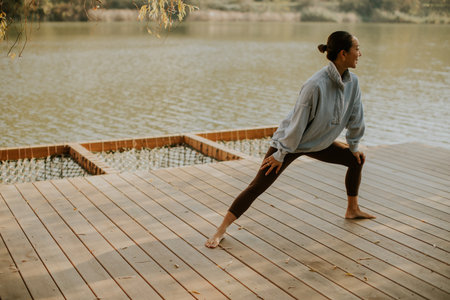 A Korean woman performs a yoga pose on a wooden deck by the serene lake, enjoying the warm sunlight and tranquil surroundings.の写真素材