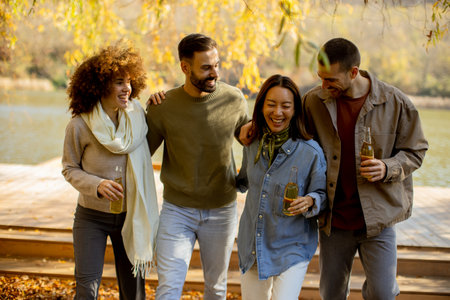 Four friends enjoy a lively moment together by the water, sharing laughter and drinks under colorful autumn leaves.の写真素材