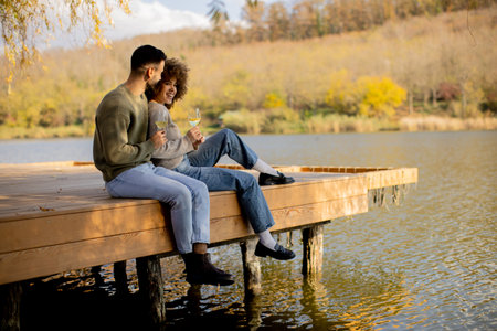 A joyful couple sits side by side on a wooden dock, sipping drinks and smiling at each other in the warmth of an autumn day.の写真素材