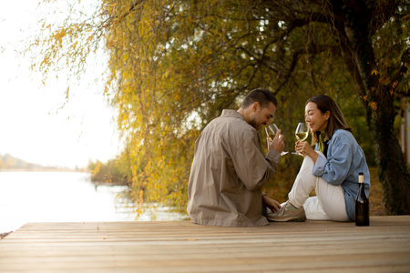 A couple shares a joyful moment by the tranquil lake at sunset, sipping wine and savoring each other's company under the trees.の写真素材