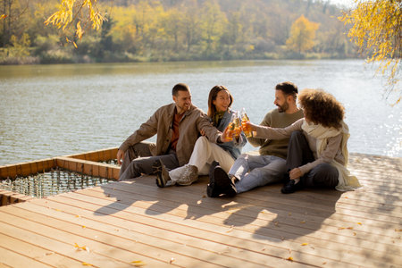 Four friends sit by the water on a wooden deck, sharing drinks and smiles on a bright autumn day.