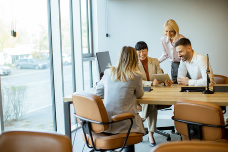 A diverse group of professionals collaborates at a conference table. They use a tablet to share ideas. The atmosphere is bright with sunlight streaming through large windows.の写真素材