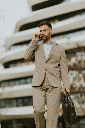 A businessman dressed in a light suit walks confidently while talking on his phone. He carries a black briefcase and is in front of a contemporary building with curved architecture.の写真素材