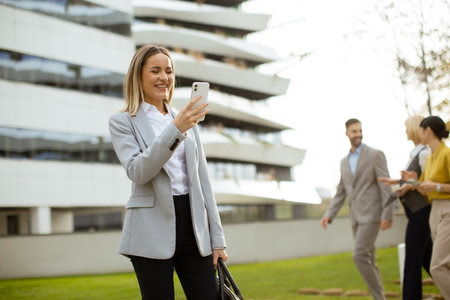 A woman stands outdoors in a stylish suit, happily looking at her phone. Two colleagues walk in the background near a contemporary building with large windows, enjoying their time.の写真素材