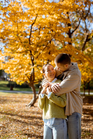 In a beautiful park, a Korean woman and a Caucasian man share a joyful moment, embracing warmly among the golden autumn leaves.の写真素材
