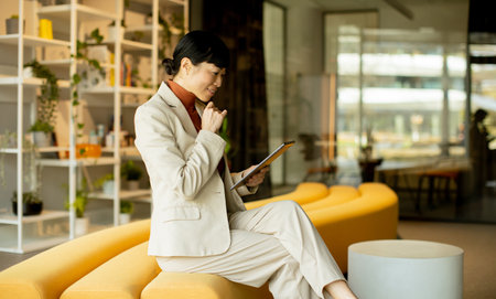In a bright, modern office, a woman dressed in a light suit sits comfortably on a yellow couch, focused on her tablet while thoughtfully resting her chin on her hand.の写真素材