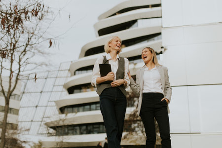 Two women are strolling down a city street in a modern architectural area. They are smiling and seem to enjoy each other's company during a pleasant afternoon walk.の写真素材