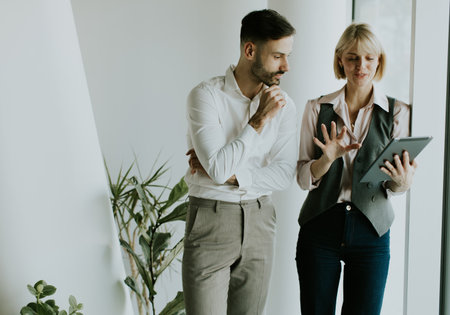 Two colleagues are engaged in conversation, examining a tablet in a bright and contemporary office. The man looks thoughtful while the woman gestures with excitement, surrounded by greenery.の写真素材