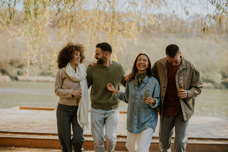 A close-knit group of friends celebrates by the lake, laughing and sharing drinks under a sunny sky, enjoying nature together.の写真素材