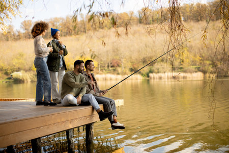Four friends gather on a wooden dock by a peaceful lake, engaging in fishing and sharing laughs. The...