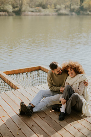 A couple is sitting on a wooden dock by a calm lake, sharing smiles and laughter while the sun shines bright.の写真素材