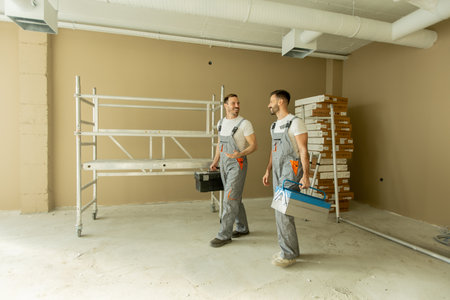 Two construction workers are walking through a newly renovated room. They carry toolboxes and...
