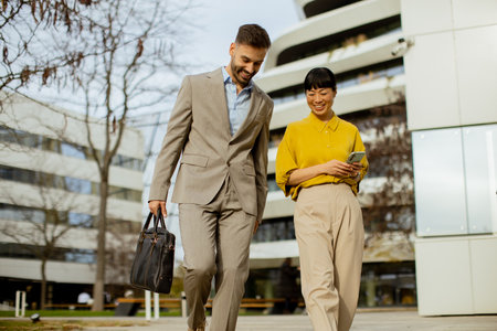 A man in a light suit carries a bag while a woman in a yellow blouse checks her phone as they stroll. They enjoy a sunny day near a contemporary architectural setting.の写真素材