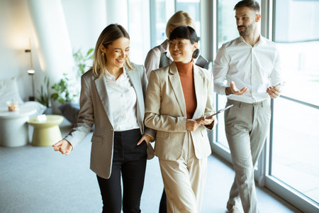 Four business professionals are engaging in a lively discussion while walking together in a well-lit office. Their attire is formal, and the atmosphere appears collaborative and positive.の写真素材