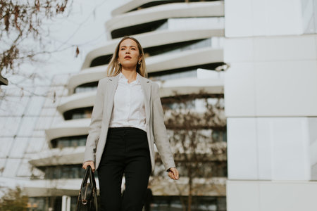 A confident business professional walks through an urban area with modern architecture. She wears a stylish outfit, showcasing a mix of professionalism and contemporary fashion during daytime.の写真素材