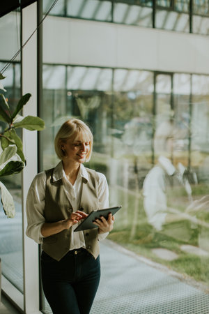 A young woman with blond hair smiles while using a tablet in a bright office. Large windows provide natural light and a view of greenery outside, creating a serene working environment.の写真素材
