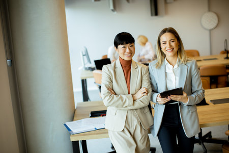 Two women stand together in a modern office, smiling and holding a tablet. They appear engaged in conversation, while colleagues work in the background.の写真素材