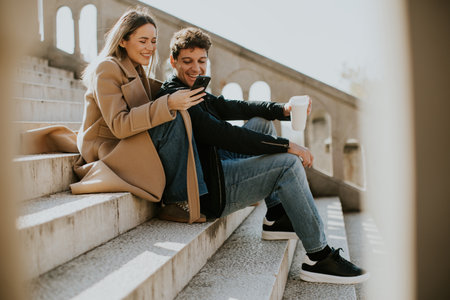 A young couple sits on stone stairs, sharing a light moment as they look at a smartphone. One holds a takeaway coffee cup, and they both seem engaged and happy in the sunlit setting.の写真素材