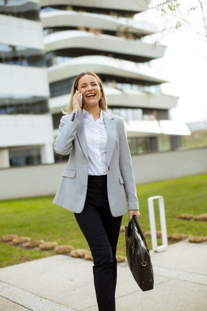 A woman dressed in a business suit walks with a smile while talking on her phone. She carries a briefcase and is near a modern building on a sunny day.の写真素材