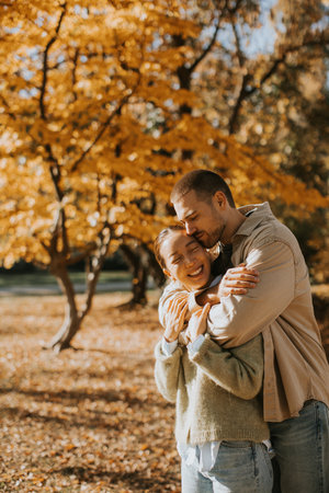 A woman smiles widely while being hugged by a man amidst vibrant autumn leaves in a sunlit park. They enjoy a joyful moment together.の写真素材