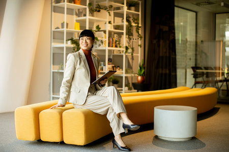 A woman in a light suit smiles while sitting on a yellow couch in a stylish office. She holds a tablet and appears engaged, surrounded by plants and shelves filled with books.の写真素材