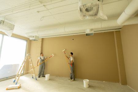 Two workers are using paint rollers to apply brown paint on a wall inside an office. A ladder and buckets are nearby, and sunlight shines in through large windows.の写真素材