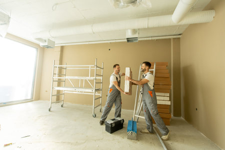Two construction workers are working together in a spacious office. They are handling building materials while scaffolding is set up in the background. Sunlight floods the room.の写真素材