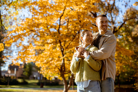 A Korean woman and Caucasian man share joyful moments in a sunny park filled with vibrant autumn leaves.の写真素材