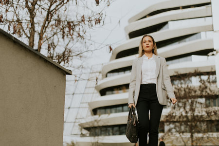 A woman dressed in a light gray suit walks through an urban area, showcasing her confidence. Behind her, striking modern architecture creates a dynamic backdrop on a clear day.の写真素材