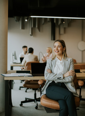 A woman sits confidently with crossed arms in a stylish office space. Behind her, colleagues engage in discussions at desks, showcasing a vibrant workplace atmosphere.の写真素材