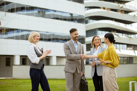 A group of four business professionals engages in a friendly handshake outside a sleek office building, enjoying a sunny afternoon.の写真素材
