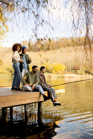 Four friends share a joyful moment while fishing from a wooden dock by a serene lake. The scene captures the beauty of autumn, with vibrant trees in the background.の写真素材