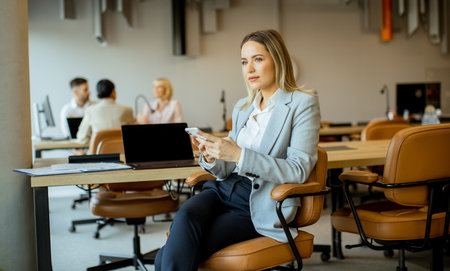 A woman in a light blue blazer sits in an office, focused on her smartphone. Other colleagues are engaged in discussions at desks in the background. The atmosphere is productive and modern.の写真素材