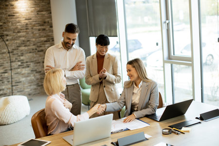 Two professionals shake hands in a bright office. A group nearby shows excitement and support for the successful agreement. This scene captures teamwork and achievement in a collaborative environment.の写真素材