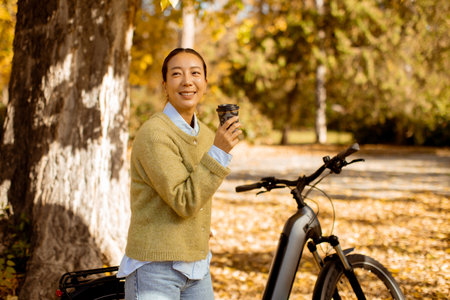 In a vibrant autumn park, a Korean woman smiles while holding a warm drink beside her bicycle, surrounded by colorful leaves.の写真素材