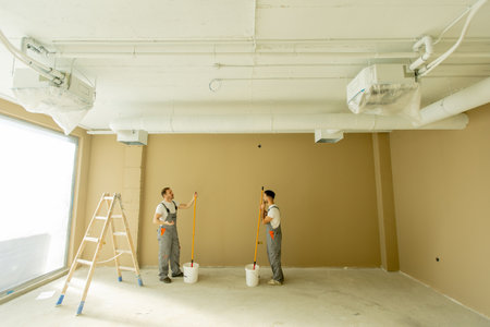 Two painters are focused on painting a wall beige in a well-lit room. They are using long rollers and standing on the floor next to a ladder and paint buckets, creating an inviting space.の写真素材