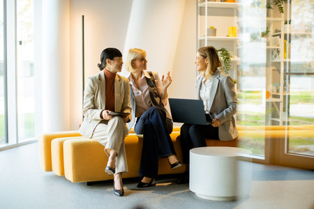 Three businesswomen engage in a lively discussion in a contemporary office lounge. They are seated on a cozy bench, sharing ideas and looking at a laptop, reflecting collaboration and teamwork.の写真素材
