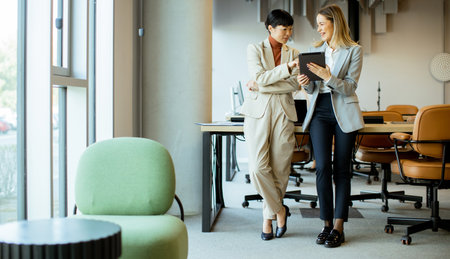 Two professional women engage in a discussion in a sleek office space, sharing ideas over a digital...