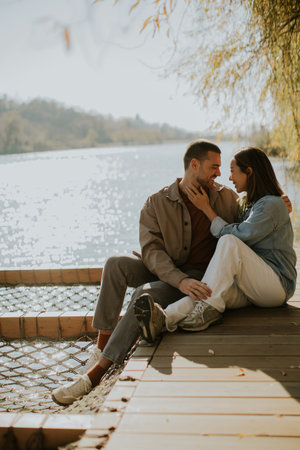 Two people sit closely on a dock, smiling and sharing a tender moment amidst sparkling water and soft sunlight.の写真素材