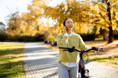 smiling young woman with bicycle talking on smartphone in park at autumnの写真素材