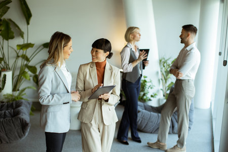 Two women talk and look at a tablet in a bright office. Two others stand nearby, engaging in...