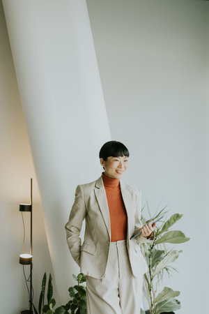 A woman in a light suit and orange turtleneck stands confidently in a modern office with plants and soft lighting, smiling while looking at her phone.の写真素材