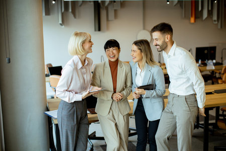 Four colleagues stand in a bright office area, sharing smiles and discussing ideas. They hold notebooks and tablets, engaging in conversation. The setting has a mix of wooden and industrial elements.の写真素材