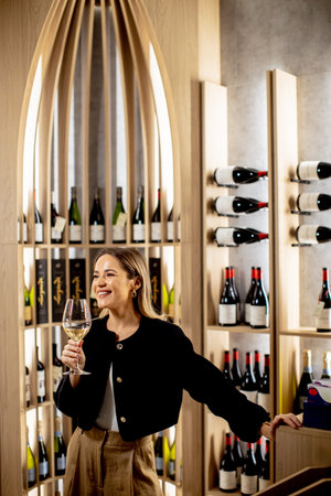 A smiling woman enjoys a glass of wine while surrounded by elegantly arranged bottles in a stylish...