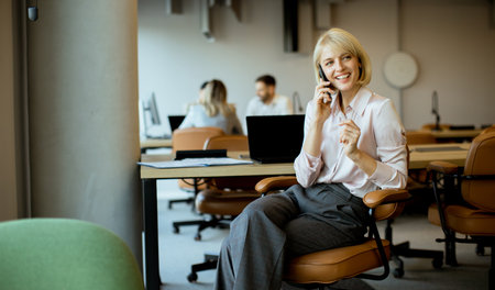 A woman sits in a chair and talks on the phone in a modern office. Colleagues are at desks in the...