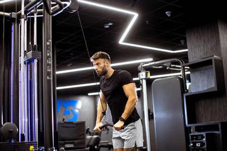 A focused man exercises on a cable machine in a stylish gym, surrounded by sleek fitness equipment...